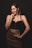 A woman stands confidently with a hand on her hip and the other hand near her chin. She wears a black crop top and a brown leather skirt. Her makeup is bold, and she has a strong, confident expression.