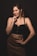 A woman stands confidently with a hand on her hip and the other hand near her chin. She wears a black crop top and a brown leather skirt. Her makeup is bold, and she has a strong, confident expression.
