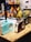 Three retro-style radios with a vintage design are displayed on a wooden table. The radios are in pastel blue, brown, and black. In the background, various colorful bags hang on a grid wall, and some printed materials like newspapers are visible. The setting appears to be a trendy shop or market.