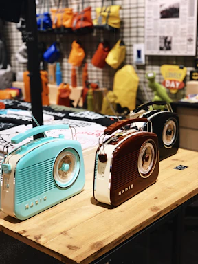 Three retro-style radios with a vintage design are displayed on a wooden table. The radios are in pastel blue, brown, and black. In the background, various colorful bags hang on a grid wall, and some printed materials like newspapers are visible. The setting appears to be a trendy shop or market.