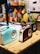 Three retro-style radios with a vintage design are displayed on a wooden table. The radios are in pastel blue, brown, and black. In the background, various colorful bags hang on a grid wall, and some printed materials like newspapers are visible. The setting appears to be a trendy shop or market.