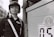 A person wearing a uniform with a helmet and sunglasses stands confidently next to a large sign or panel. The uniform includes distinctive badges and insignia. The setting appears urban with a glimpse of buildings in the background. The image is in black and white.