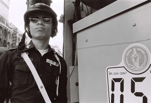 A person wearing a uniform with a helmet and sunglasses stands confidently next to a large sign or panel. The uniform includes distinctive badges and insignia. The setting appears urban with a glimpse of buildings in the background. The image is in black and white.
