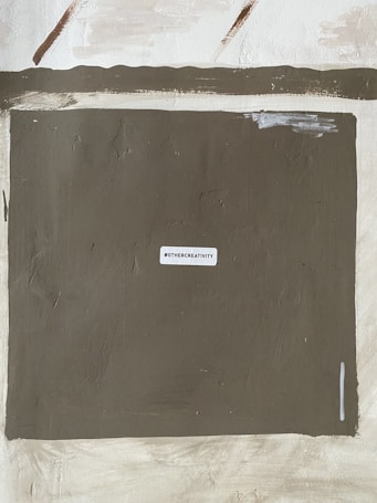 A textured wall with a large, dark rectangular area in the center. A small white label with the text '#OTHERCREATIVITY' is affixed centrally on this dark section. The surrounding wall areas are painted in lighter, varied shades of beige and brown, creating a rustic appearance.