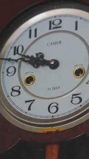 A vintage clock face with Roman numerals encircling the dial. The hands of the clock are ornate in design, and the clock appears to be a 31-day model, indicated by the text. The name 'CAMER' is visible at the top of the clock face. The frame of the clock appears to be wooden with a visible area of slight rust beneath the dial.