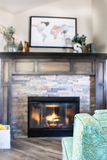 A cozy living room setting with a stone fireplace, above which hangs a framed world map. Decorative items such as books and vases are placed on the wooden mantel. A soft, light-blue patterned chair is in the foreground, facing the warm glow of the fire. Wooden flooring adds a rustic charm.