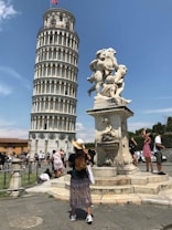 A large, distinctive leaning tower is in the background, with a marble statue featuring cherubic figures in the foreground. Numerous tourists are present, some taking photos and others appearing to be posing. A person with long hair wearing a hat and a long skirt stands near the statue.