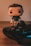 A pop culture figurine with a stylized design stands next to a video game controller on a wooden surface. The figurine has a cute, cartoonish appearance with an oversized head and minimal facial features. The game controller is a modern design with colorful buttons and analog sticks.