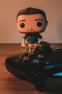 A pop culture figurine with a stylized design stands next to a video game controller on a wooden surface. The figurine has a cute, cartoonish appearance with an oversized head and minimal facial features. The game controller is a modern design with colorful buttons and analog sticks.