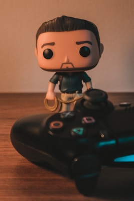 A pop culture figurine with a stylized design stands next to a video game controller on a wooden surface. The figurine has a cute, cartoonish appearance with an oversized head and minimal facial features. The game controller is a modern design with colorful buttons and analog sticks.
