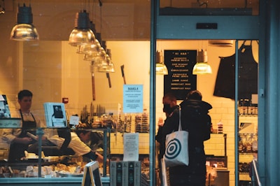 A cozy deli shop viewed from the outside, featuring several people inside. One is a worker behind the counter arranging goods, while a couple of customers stand in line, one holding a branded tote bag. The interior is warmly lit with hanging industrial-style lamps, and the counter displays various food items. The walls are decorated with menus or signs, and there is an apron hanging nearby.