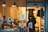 A cozy deli shop viewed from the outside, featuring several people inside. One is a worker behind the counter arranging goods, while a couple of customers stand in line, one holding a branded tote bag. The interior is warmly lit with hanging industrial-style lamps, and the counter displays various food items. The walls are decorated with menus or signs, and there is an apron hanging nearby.