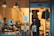 A cozy deli shop viewed from the outside, featuring several people inside. One is a worker behind the counter arranging goods, while a couple of customers stand in line, one holding a branded tote bag. The interior is warmly lit with hanging industrial-style lamps, and the counter displays various food items. The walls are decorated with menus or signs, and there is an apron hanging nearby.