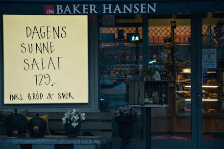 A storefront with a large illuminated sign advertising a daily healthy salad for 129 currency units, including bread and butter. The window display and entrance are visible, featuring a rustic crate with items and decorative elements such as flowers and lanterns.
