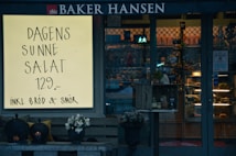 A storefront with a large illuminated sign advertising a daily healthy salad for 129 currency units, including bread and butter. The window display and entrance are visible, featuring a rustic crate with items and decorative elements such as flowers and lanterns.
