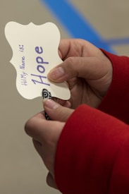 A pair of hands holding a paper cut-out with a decorative edge. The paper has handwritten text in purple that reads 'Hi! My Name is: Hope'. The fingers are partially visible and the sleeve of the person is red.