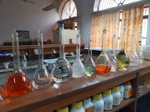 A laboratory setting features a wooden countertop lined with a series of volumetric flasks containing liquids of various colors, such as clear, orange, and green. Behind the flasks, there are shelves with chemical containers, and windows with patterned yellow curtains allow natural light to enter. The setting suggests an environment focused on scientific research or experimentation.