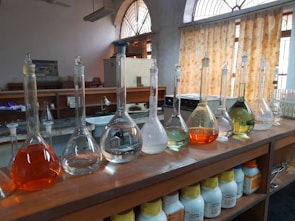 A laboratory setting features a wooden countertop lined with a series of volumetric flasks containing liquids of various colors, such as clear, orange, and green. Behind the flasks, there are shelves with chemical containers, and windows with patterned yellow curtains allow natural light to enter. The setting suggests an environment focused on scientific research or experimentation.