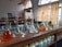 A laboratory setting features a wooden countertop lined with a series of volumetric flasks containing liquids of various colors, such as clear, orange, and green. Behind the flasks, there are shelves with chemical containers, and windows with patterned yellow curtains allow natural light to enter. The setting suggests an environment focused on scientific research or experimentation.