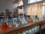 A laboratory setting features a wooden countertop lined with a series of volumetric flasks containing liquids of various colors, such as clear, orange, and green. Behind the flasks, there are shelves with chemical containers, and windows with patterned yellow curtains allow natural light to enter. The setting suggests an environment focused on scientific research or experimentation.