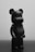 A matte black figurine that resembles a bear stands on a reflective surface, showing minimalist design features. The lighting creates soft shadows and highlights on the smooth surface, giving it a sleek appearance.
