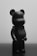 A matte black figurine that resembles a bear stands on a reflective surface, showing minimalist design features. The lighting creates soft shadows and highlights on the smooth surface, giving it a sleek appearance.