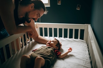 A father leans over the side of a crib, smiling and interacting with his baby, who is lying on a mattress. The baby is wearing a red and brown outfit and looking up at the father. The room is dimly lit, with walls adorned with a few small, square photo frames.