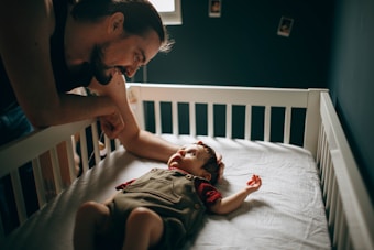 A father leans over the side of a crib, smiling and interacting with his baby, who is lying on a mattress. The baby is wearing a red and brown outfit and looking up at the father. The room is dimly lit, with walls adorned with a few small, square photo frames.