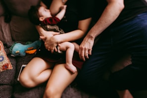 A nurturing scene featuring an adult holding a baby closely. The adult and baby are seated on a couch, surrounded by a cozy environment. There is a colorful stuffed toy and a patterned pillow nearby, adding warmth to the setting.