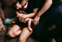 A nurturing scene featuring an adult holding a baby closely. The adult and baby are seated on a couch, surrounded by a cozy environment. There is a colorful stuffed toy and a patterned pillow nearby, adding warmth to the setting.
