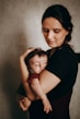 A woman gently cradles a sleeping baby in her arms, both appearing peaceful. The setting has a soft, muted color palette, adding warmth to the intimate scene. The woman’s serene expression and careful hold suggest tenderness and protection.