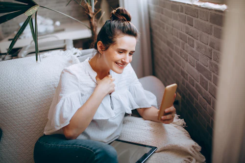 Une jeune femme assise sur un canapé blanc sourit en regardant son téléphone, une tablette posée sur les genoux