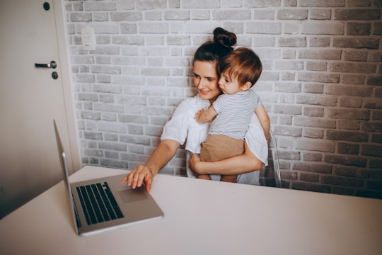 A woman with dark hair is sitting at a desk holding a toddler with light brown hair. They are both looking at a laptop on the table, which is situated against a light gray brick wall. The woman is casually dressed in a white shirt, and the child is wearing a striped shirt and brown shorts.