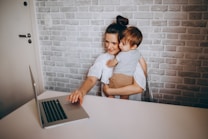 A woman with dark hair is sitting at a desk holding a toddler with light brown hair. They are both looking at a laptop on the table, which is situated against a light gray brick wall. The woman is casually dressed in a white shirt, and the child is wearing a striped shirt and brown shorts.