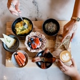 A wooden board holds an assortment of breakfast foods, including a bowl of scrambled eggs garnished with greens, a bowl of grains with seeds, a plate with a waffle topped with berries and a strawberry, and slices of triangular grilled bread. To the side, there is a bowl with orange slices and another with bacon strips. A small latte with a heart-shaped design in the foam is also present. Two hands are visible, one holding the latte and the other reaching for the bread.