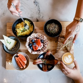 A wooden board holds an assortment of breakfast foods, including a bowl of scrambled eggs garnished with greens, a bowl of grains with seeds, a plate with a waffle topped with berries and a strawberry, and slices of triangular grilled bread. To the side, there is a bowl with orange slices and another with bacon strips. A small latte with a heart-shaped design in the foam is also present. Two hands are visible, one holding the latte and the other reaching for the bread.