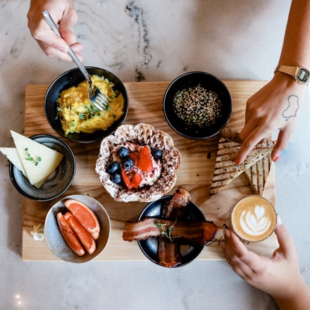 A wooden board holds an assortment of breakfast foods, including a bowl of scrambled eggs garnished with greens, a bowl of grains with seeds, a plate with a waffle topped with berries and a strawberry, and slices of triangular grilled bread. To the side, there is a bowl with orange slices and another with bacon strips. A small latte with a heart-shaped design in the foam is also present. Two hands are visible, one holding the latte and the other reaching for the bread.