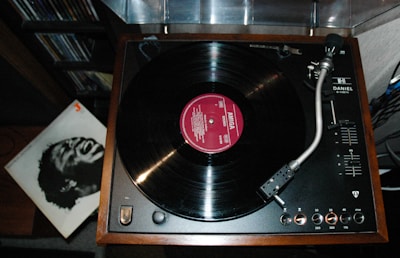 A vintage turntable with a vinyl record playing, featuring a wooden frame and various control knobs. To the side, there is a vinyl cover with a black and white image of a person expressing strong emotion.