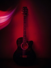 An acoustic guitar is positioned against a dark background, with a red light casting dramatic shadows and highlights. The guitar's strings and tuning pegs are clearly visible, while the stylized light effect on the left adds a sense of dynamic motion.