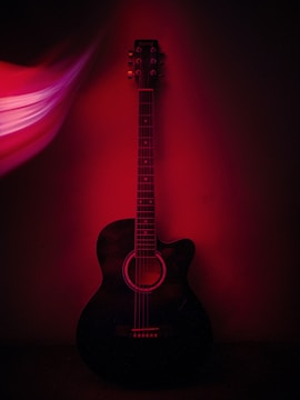 An acoustic guitar is positioned against a dark background, with a red light casting dramatic shadows and highlights. The guitar's strings and tuning pegs are clearly visible, while the stylized light effect on the left adds a sense of dynamic motion.
