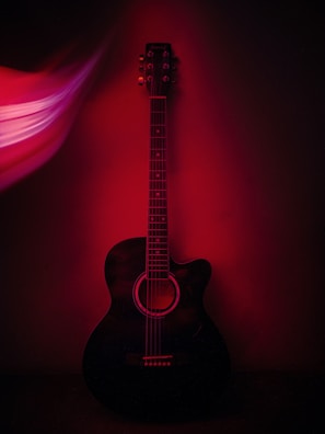 An acoustic guitar is positioned against a dark background, with a red light casting dramatic shadows and highlights. The guitar's strings and tuning pegs are clearly visible, while the stylized light effect on the left adds a sense of dynamic motion.