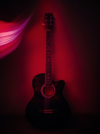 An acoustic guitar is positioned against a dark background, with a red light casting dramatic shadows and highlights. The guitar's strings and tuning pegs are clearly visible, while the stylized light effect on the left adds a sense of dynamic motion.