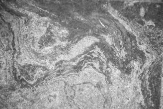 A textured surface with swirling patterns reminiscent of natural stone or marble. The image is monochromatic, featuring variations of gray tones with lighter and darker areas blending together.
