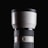 A large, white telephoto camera lens with a black cap, featuring fine ribbed texture on the adjustable rings. The brand logo is displayed on the upper part of the lens. The background is a solid black, highlighting the sleek and professional design of the lens.