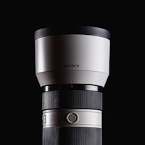 A large, white telephoto camera lens with a black cap, featuring fine ribbed texture on the adjustable rings. The brand logo is displayed on the upper part of the lens. The background is a solid black, highlighting the sleek and professional design of the lens.