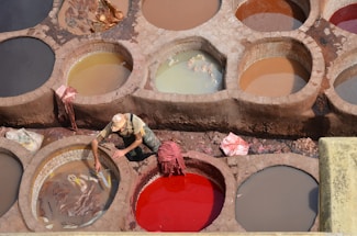A worker is tending to large round vats filled with different colored liquids used in the leather tanning process. The earthy tones and varied shades create a mosaic of circular patterns. The worker wears a hat and casual clothing, focusing on their task. Some vats contain animal hides soaking in the mixture.