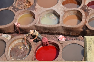 A worker is tending to large round vats filled with different colored liquids used in the leather tanning process. The earthy tones and varied shades create a mosaic of circular patterns. The worker wears a hat and casual clothing, focusing on their task. Some vats contain animal hides soaking in the mixture.