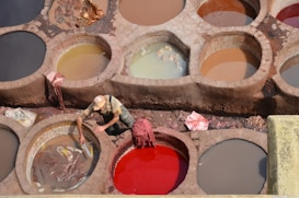 A worker is tending to large round vats filled with different colored liquids used in the leather tanning process. The earthy tones and varied shades create a mosaic of circular patterns. The worker wears a hat and casual clothing, focusing on their task. Some vats contain animal hides soaking in the mixture.