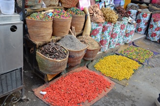 Various baskets and containers filled with colorful dried goods and spices are displayed at an outdoor market stall. The items range in color, including green, red, yellow, and natural earth tones. The baskets are made of woven materials, and some floral-patterned bags are also visible. Brightly colored powders or seeds are spread out on mats on the ground.