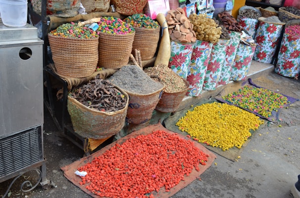 Various baskets and containers filled with colorful dried goods and spices are displayed at an outdoor market stall. The items range in color, including green, red, yellow, and natural earth tones. The baskets are made of woven materials, and some floral-patterned bags are also visible. Brightly colored powders or seeds are spread out on mats on the ground.